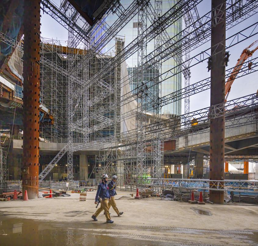 Construction workers walk through the Takanawa Gateway City site, surrounded by scaffolding, steel beams, and building materials.