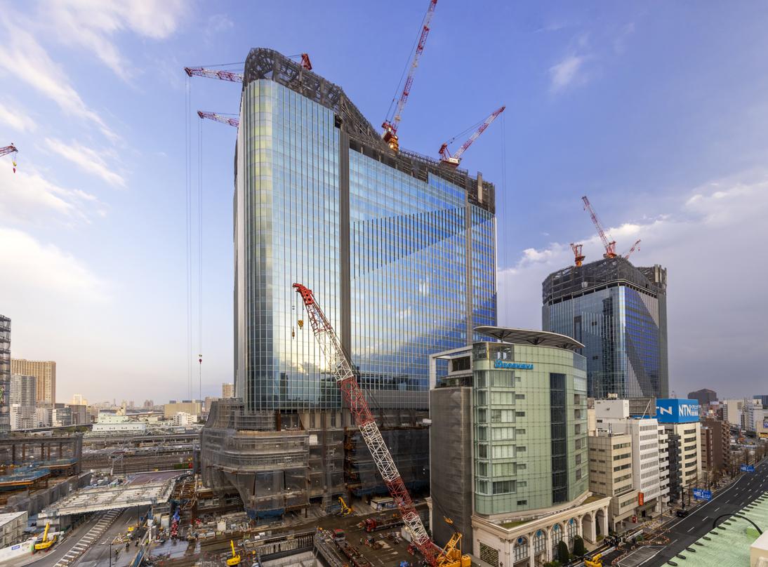 The Takanawa Gateway City building under construction, with cranes surrounding the glass skyscraper in an urban setting.
