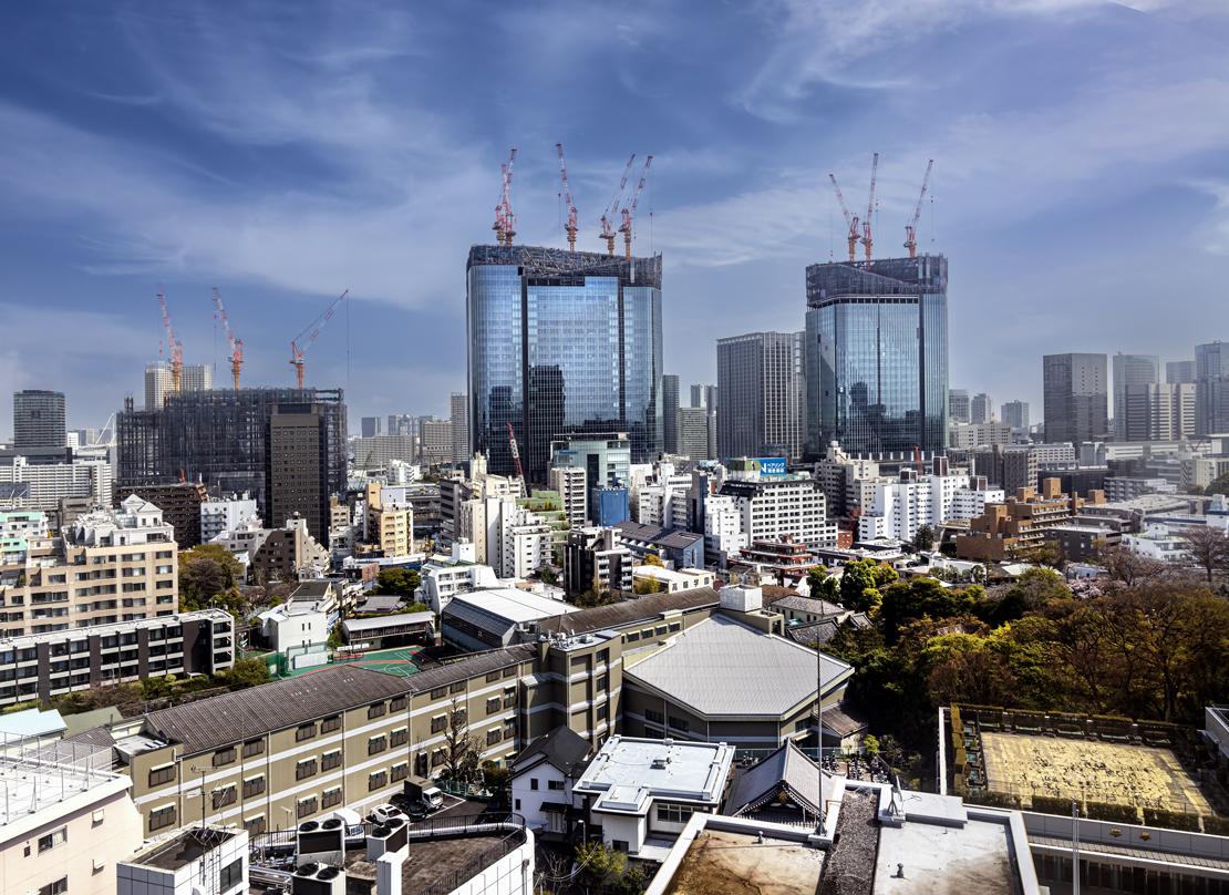 Takanawa Gateway City skyscrapers under construction, with cranes atop the buildings, set against a cityscape with a mix of modern and traditional structures.