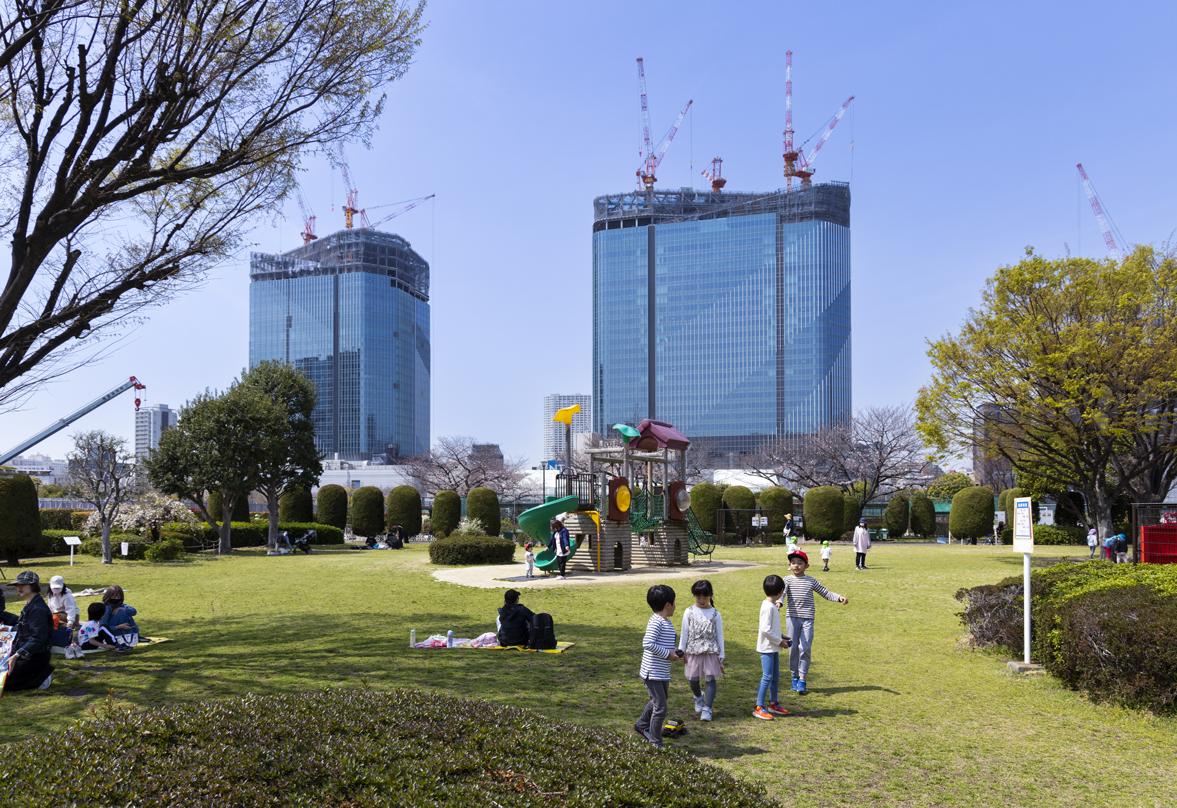 Children play in a park with the Takanawa Gateway City skyscrapers under construction in the background, featuring cranes and glass facades.