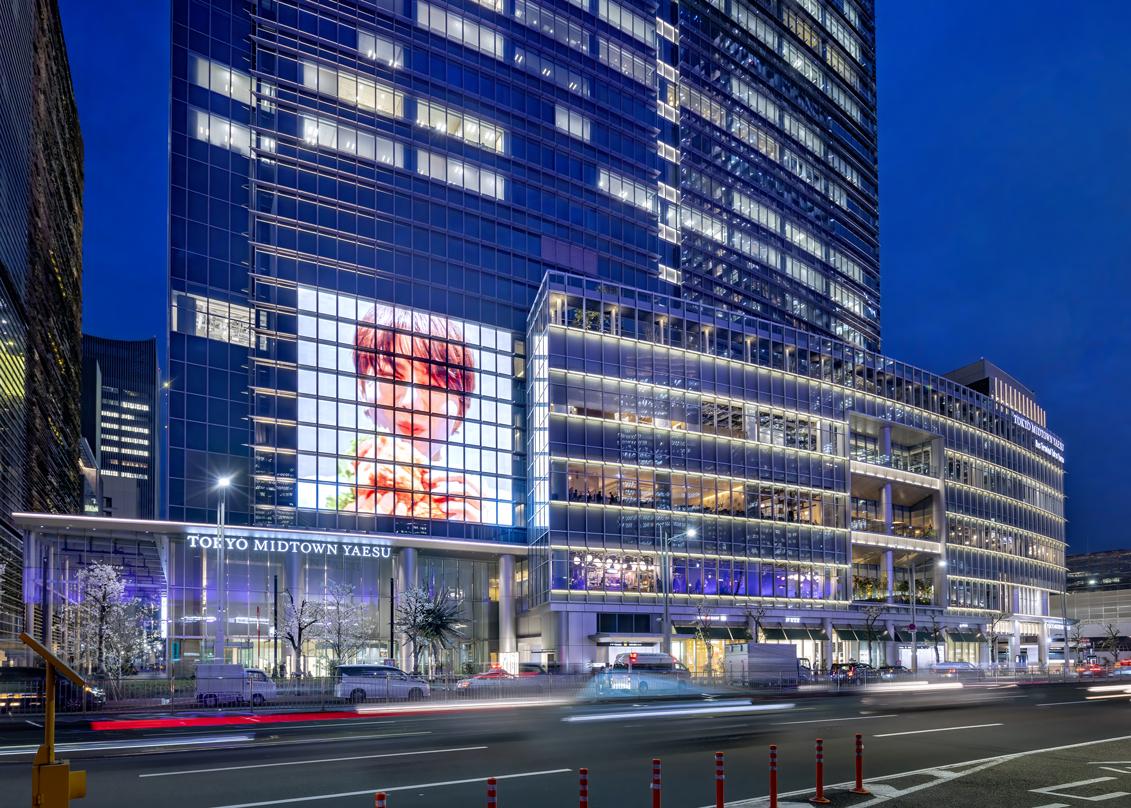 Tokyo Midtown Yaesu at night, with illuminated glass facades and a large digital billboard displaying an image. Cars and pedestrians are visible on the street in front.