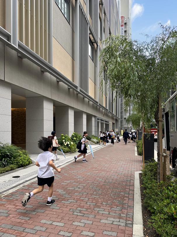 Elementary school children play along a landscaped pedestrian walkway next to a school at Tokyo Midtown Yaesu, featuring greenery.
