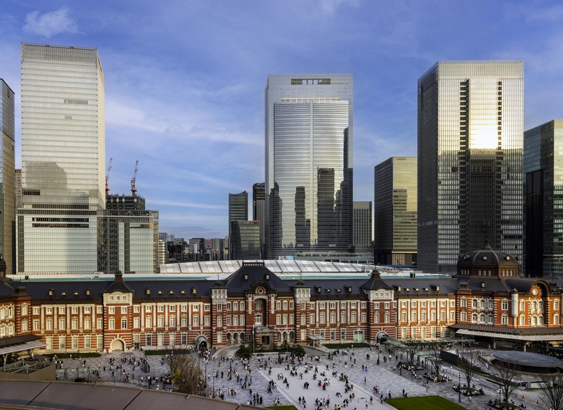 Tokyo Station with its historic red-brick facade, bustling with people in the foreground and modern skyscrapers in the background.