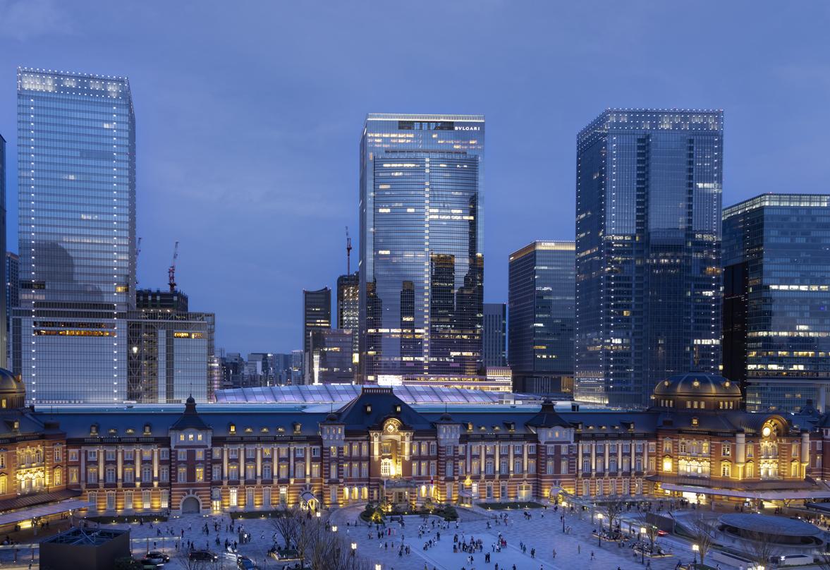 Tokyo Station at dusk with its historic red-brick facade, bustling with people, and Tokyo Midtown Yaesu skyscrapers illuminated in the background.
