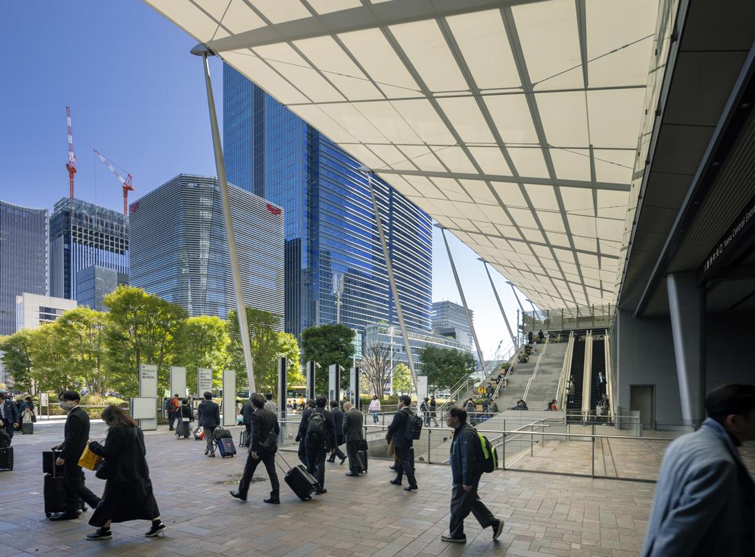 People walking under a modern canopy at Tokyo Midtown Yaesu, with glass skyscrapers and greenery in the background on a sunny day.